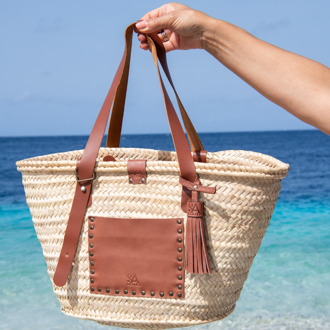 Handgemaakte en gepersonaliseerde rieten tas met leren vakken en een leren lus aan de buiten en binnenzijde gefotografeerd aan de hand aan zee