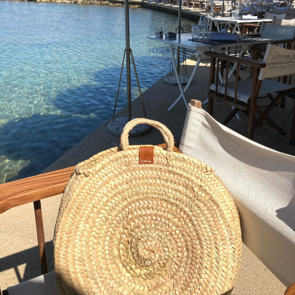 Ronde rieten tas met raffia handvatten op een stoel op een terras aan zee